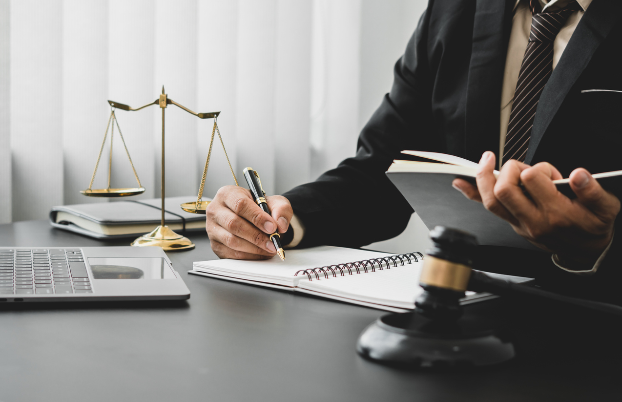 Lawyer in suit writing notes with pen, scales of justice and gavel on desk, emphasizing legal expertise in criminal law.