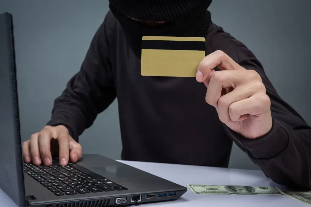 Person in black attire holding a credit card in front of a laptop, representing credit card fraud, with cash visible on the table, relevant to legal defense services in Tomball.