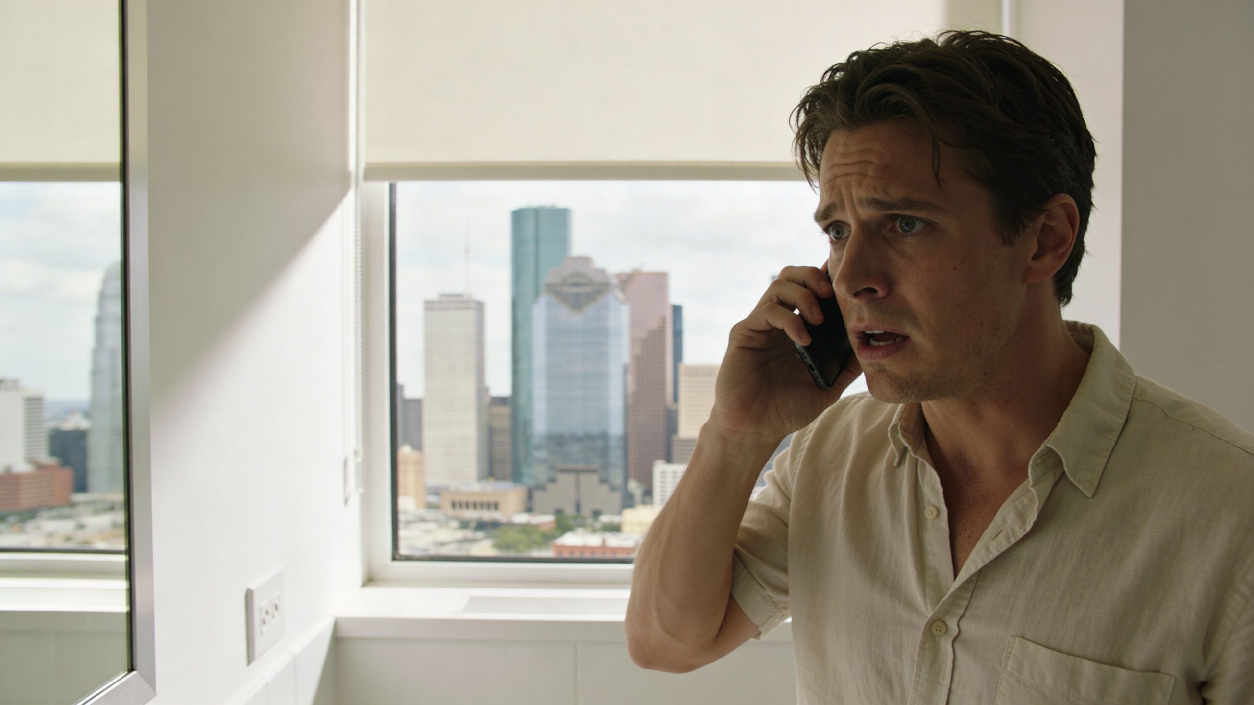 A concerned man talks on his smartphone while standing in front of a Houston skyline window.