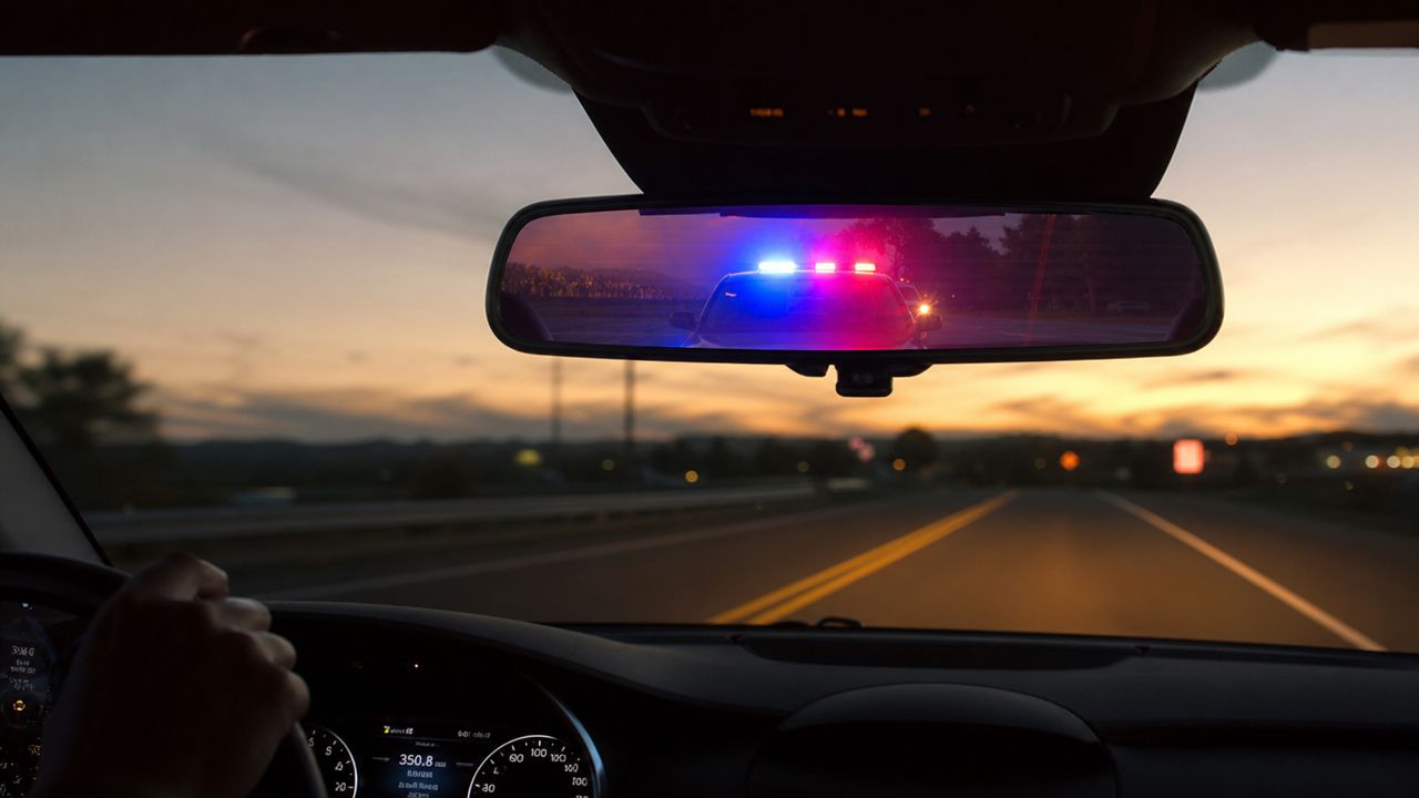 A view from inside a car looking into the rearview mirror at a police cruiser flashing blue lights.